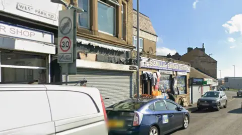 Cars and vans parked up at the side of a road in front of a row of shops.