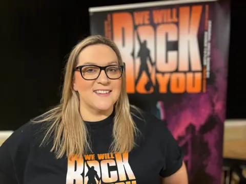 A blonde haired woman with glasses, wearing a black t-shirt, smiles at the camera