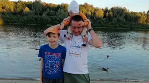 Anton Honchanka Anton in a white t shirt stood with two boys. One is on his shoulders holding onto his head. The other is standing by his side with a blue t shirt on.