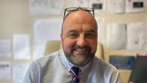 Shows a man with a beard and receding hairline with dark-rimmed glasses on the top of his head. He is wearing a blue button-down shirt with a blue and pink striped tie and is sitting in a classroom. 
