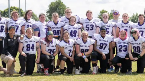 Peterborough Royals A team photo pf the Peterborough Royals - all wearing a white and purple kit, smiling for camera. Some are standing in the back row while the front row people are kneeling down. Coach Maurice is also kneeling down in the left in the front row.