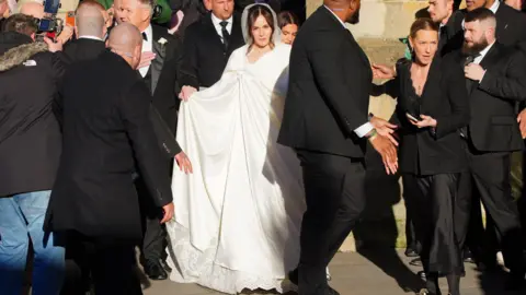 PA Media Holly Ramsay entering the church surrounded by security staff. She is wearing a white cape over her white wedding dress. 