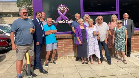 A group of people pose for a photo outside Charlie's Cafe, a building with green and purple exterior accents. The cafe's signage displays a steaming coffee cup logo alongside the name 'Charlie's Cafe' in white lettering. The individuals, dressed in a mix of casual and semi-formal clothing, stand together on a bright, sunny day, suggesting a celebratory or community-focused occasion