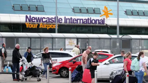 People with luggage standing outside Newcastle Airport near to parked cars. A sign on the building has purple and yellow writing which reads: "Newcastle International Your Airport". There is a long shelter, like a bus stop, in front of the airport terminal.