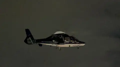 MICH2255 A dark-coloured helicopter seen flying in the night sky, with a white section beneath and also just below the rotor blades. 