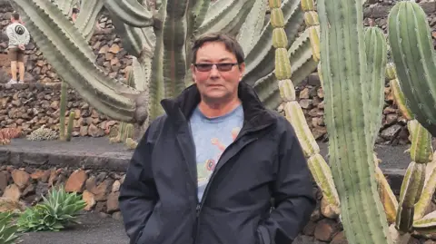 Family handout Frank Ingram is standing in front of several large cacti in a landscaped area outside. He is wearing a black jacket and blue T-shirt with a frog on it. He is wearing lightly-tinted sunglasses. 