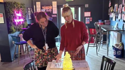 Kat Dooney An unnamed woman with brown hair in a ponytail wearing a black top and flowery skirt and black tights listens to artist Janine Walker with blond hair in plaits wearing a red jumper and a skirt with a flower pattern. Janine is talking about the maquette of the puppet which is on a table in front of the women. 