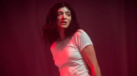 Lorde on stage at Glastonbury. The singer, lit in a red light, wears a white T-shirt, her arms by her side. Her dark hair is worn loose and messy. She looks over her left shoulder towards the crowd. 