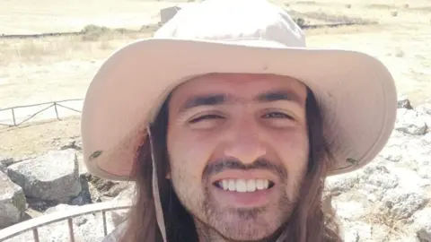 Family handout Michael Frison pictured in a desert-like area on a hot summer's day. He is wearing a cream coloured sun hat and has long brown hair and stubble. He is smiling at the camera
