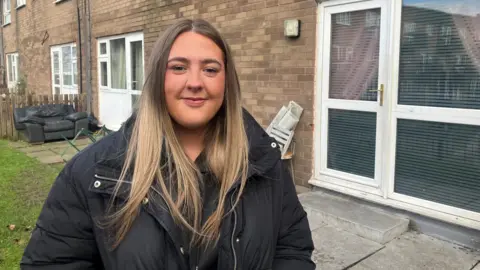 Richard Stead/BBC Kelsey Schofield, 18, who has long straight blonde hair and is wearing a thick black coat, smiles at the camera while standing on a lawn outside a ground floor flat. 