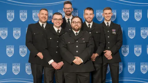 Six police officers in uniform smile and look directly at the camera. They each have their hands clasped in front of them and are all standing in front of a blue step and repeat banner with Thames Valley Police's logo on it. 