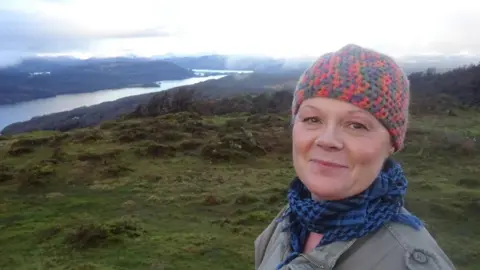 Cat, a woman wearing a red and grey woolly hat and blue scarf, she smiles as she stands in a field, you can see a large lake in the background.