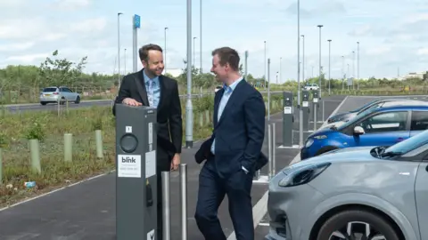 SYMCA Two men in dark suits stand next to a grey car parked in a marked bay next to an EV charging post