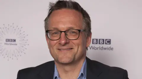 BBC/PA Media Michael Mosley, wearing a dark suit and blue shirt, smiles in a press photograph. He has short dark grey hair and is wearing black-rimmed glasses.