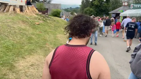 Man wearing red vest standing with back to camera, showing curly mullet hair style. He is stood on a path with a grassy verge on the left and market stalls on the right, with people walking past.