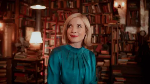 Ian Mann Lucy Worsley has shoulder-length blonde hair and wearing a turquoise outfit. She is in a room filled with old books.