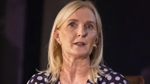 PA Media A long-blonde haired woman with a microphone on her left cheek, wears a navy dress with white polka dots. She has a small diamond earing in her left ear.