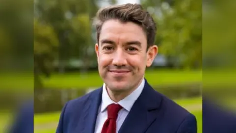 LDRS Alan Strickland, a white man with brown hair, wearing a navy blue suit and red tie, smiling