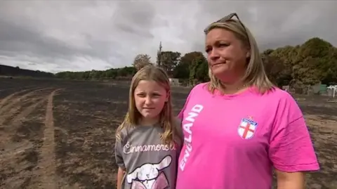 A woman with shoulder length blonde hair and sunglasses on her head is wearing a pink t-shirt that says "england" in white writing. She has her arm around her young daughter who has mousy brown hair and is wearing a brown t-shirt with a white rabbit on it. They are standing in a field that is blackened and singed by fire