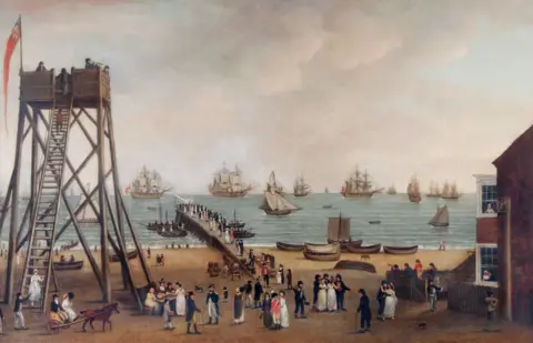 Norfolk Museums Service Painting of Great Yarmouth port and jetty in late 17th century. Many ships can be seen in the sea, with row-boats on the shore line. A look-out tower is full of people, one with a telescope, while a woman climbs down the tower from a ladder to be greeted by a waiting man. Throngs of people are on the jetty and the beach. The women wear mostly white or pale-coloured dresses. Some men wear workers clothing while others wear suits of the gentry. One man is wearing a red jacket and hat denoting he is a member of the military.