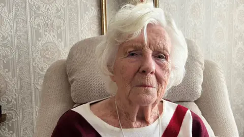 An elderly woman with white hair sat in an arm chair looking at the camera.