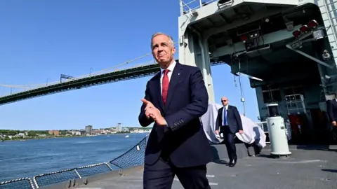 Mark Carney in a dark blue suit and red tie, walks by the water at the t HMC Dockyard