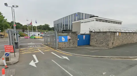 A image of the front entrance to the current Babcock building in Plymouth. It is a large glass building with a brick entrance and security gates. There are cars parked outside the front of the building.