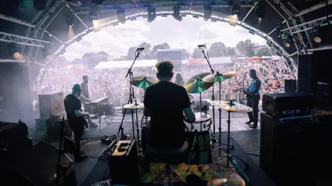 A band playing on a stage, viewed from the back, behind the drummer. In the distance is a crowd,