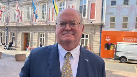 BBC Constable Simon Crowcroft is stood in front of St Helier parish hall. He is wearing a dark blue suit and white shirt.