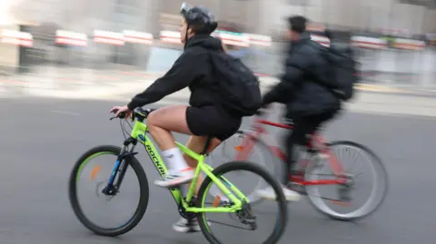 Female commute on bright yellow bicycle with helmet, rucksack,black hooide and black cycling shorts.