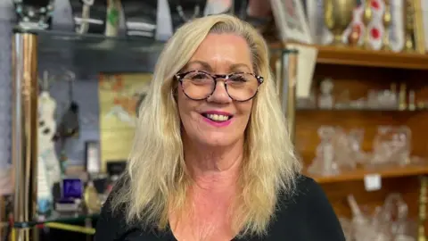 Audrey Houston smiles at the camera with items on sale in the BRICC shop in the background. She has long, blonde hair and is wearing dark glasses with pink speckles.