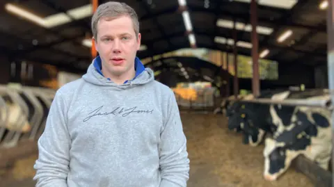 Ross Beatty looks forward, is wearing a grey jack and jones hoodie. He has grey hair and blue eyes. Behind him there is a row of black and white cows eating hay. 