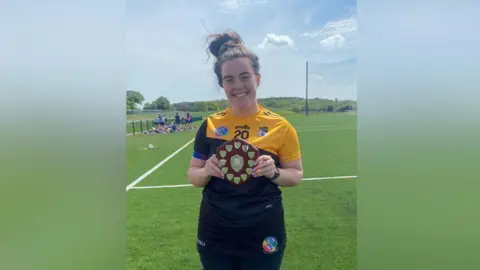 Caoihm Mallon A woman in a yellow and black GAA jersey. She is holding a brown and silver trophy.