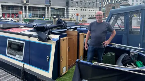 BBC/Nathan Turvey Ken Tracey standing by the doors of his blue and white narrow boat. He is bald, wearing glasses and a blue top and jeans. There are buildings in the background.