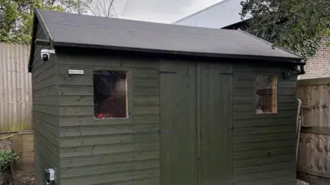Andrej Miskolci A dark green shed with a sloped roof. It is in a garden with wooden panelled fences behind it.