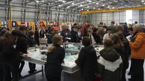 Lots of school pupils crowded around tables where are covered in pieces of Lego. They are in an aircraft hanger with bunting hung along one wall 