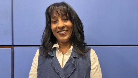 A woman with long dark hair is wearing blue and white and smiling at the camera. She is in front of a blue background.