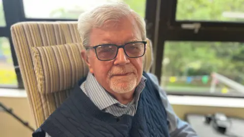 Kevin is looking at the camera and smiling. He is wearing a blue and white stripped shirt and a navy knitted tank top. He has short white hair and glasses.