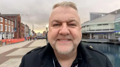 A head and shoulders image of a man standing on a dockside, with a wide promenade and converted warehouse buildings to the left and a dock and large 1980s shopping centre to the right. He has short blonde hair, swept back, stubble and is smiling. He is wearing a smart black coat.