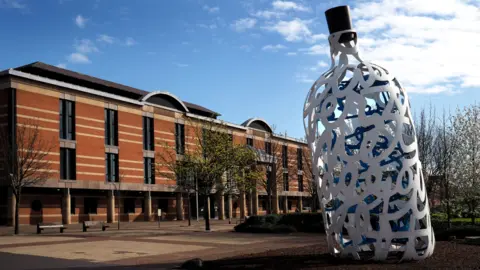 Teesside Crown Court is a long brown building of two storeys with lots of pillars over a walkway. There are a few trees in front of it - and a large white sculpture in the shape of a bottle dominates the image 