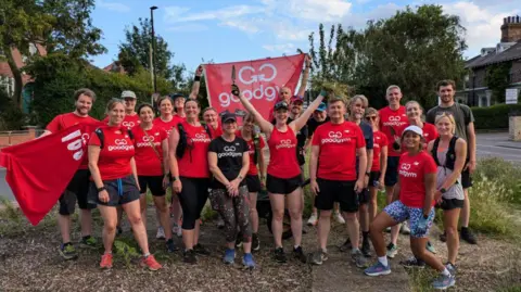 About 20 people stand on a street holding red GoodGym flags and wearing red and black T shirts with shorts and trainers.