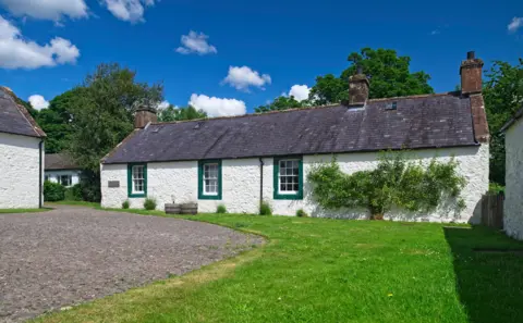 Ian Findlay An image of a farmhouse below a blue sky, with green grass and a pavement in front