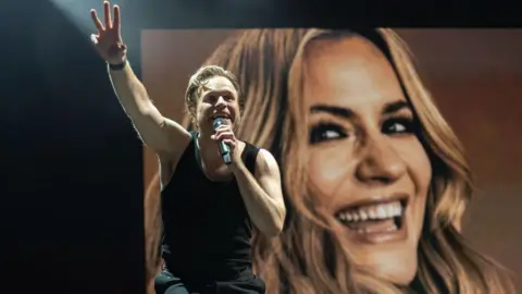 Getty Images Olly Murs, singing into a microphone and in front of a picture of Caroline Flack, performs at Flackstock 2024 at Englefield House on July 22, 2024