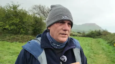 A man in a gray woolly hat is pictured looking in the direction of the camera. He is wearing a blue coat over a dark fleece. There is grass and some bushes behind him, as well as some hills in the distance. 