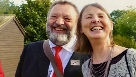 Lesley Clarke Richard Still and Lesley Clarke standing close together and smiling into the camera. Richard, who has short hair and a beard, is wearing a suit, white shirt and red tie. Lesley, who has shoulder length hair, is wearing a black top and a necklace.