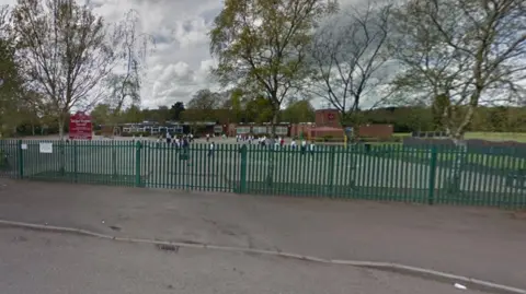 Google A generic image of Michael Drayton Junior School, taken from outside the site, which has green railings at the front, close to a red and white school sign. Children are outside the building in the distance.
