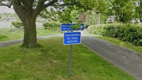 A country lane runs along the right of the picture while on the left a blue road sign reading "unsuitable for long vehicles" can be seen on a grass verge under a tree.