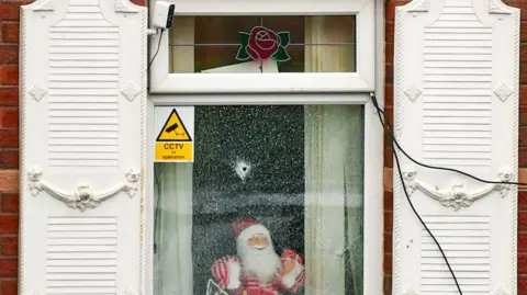 A single bullet hole is in the middle of the glass window. Either side of the windows are white shutters. There is a Santa figure on the window sill and a sticker with "CCTV in operation" placed beside it.