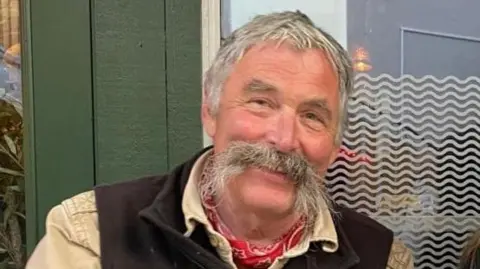 A head and shoulder photograph of David Oliver from Marlborough smiling with grey hair and a large grey moustache wearing a pale yellow shirt, a brown waistcoat and a red and white neckerchief, with a green glass door in the background. 