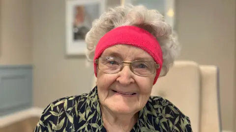 Judith Snell/BBC. Judith Snell, una señora mayor con gafas que lleva una diadema rosa brillante. En la foto aparece sonriendo.